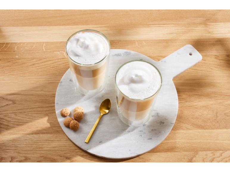 Deux verres de latte macchiato en couches avec de la mousse, une cuillère dorée et des biscuits sur une planche en marbre.