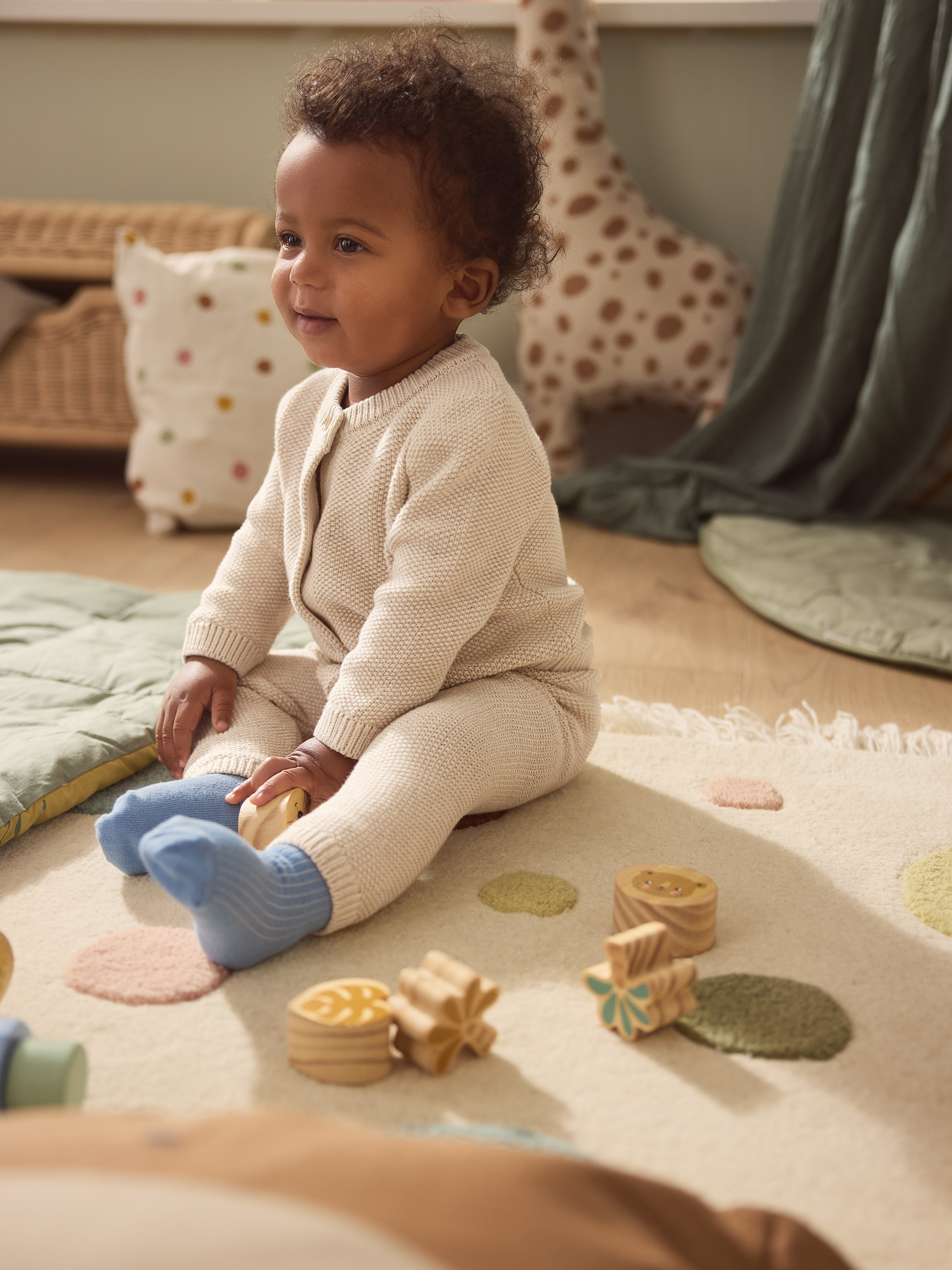 Bébé souriant en grenouillère tricotée beige, jouant avec des blocs en bois sur un tapis.