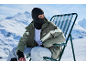 Homme en cagoule et veste de ski verte assis sur une chaise dans les montagnes enneigées.