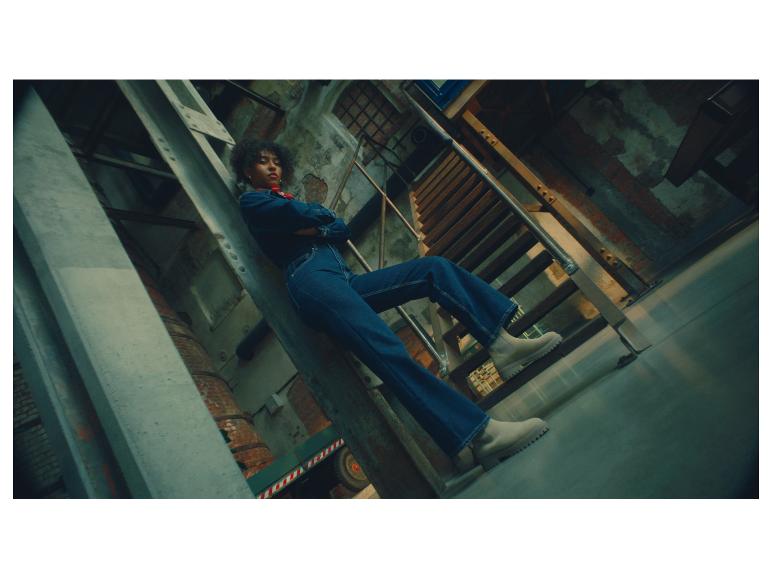 Une femme en combinaison en jean et bottes claires pose sur des escaliers industriels.