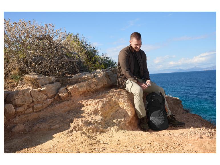 Homme en tenue de randonnée assis sur un rocher au bord de la mer avec un sac à dos.
