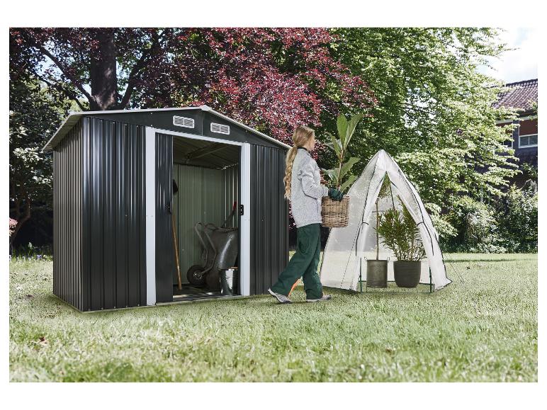 Une femme jardine à côté d'un abri de jardin et d'une petite serre dans la cour.
