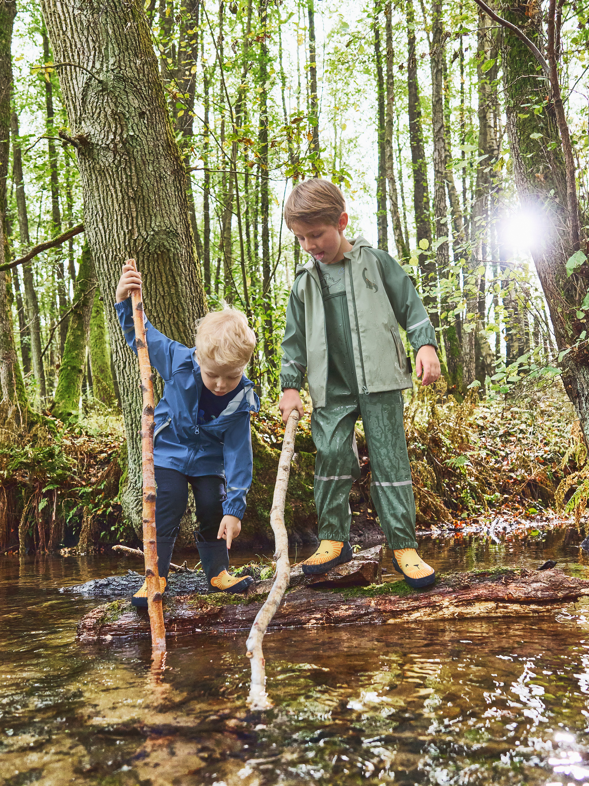 Deux enfants en cirés et bottes de pluie jouent avec des bâtons dans un ruisseau en forêt.