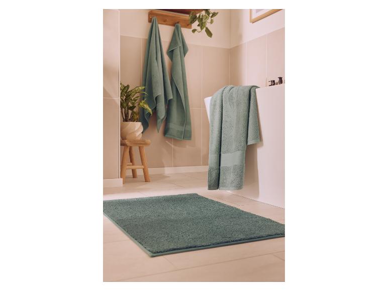 Salle de bain avec serviettes et tapis verts, une plante et un tabouret en bois.