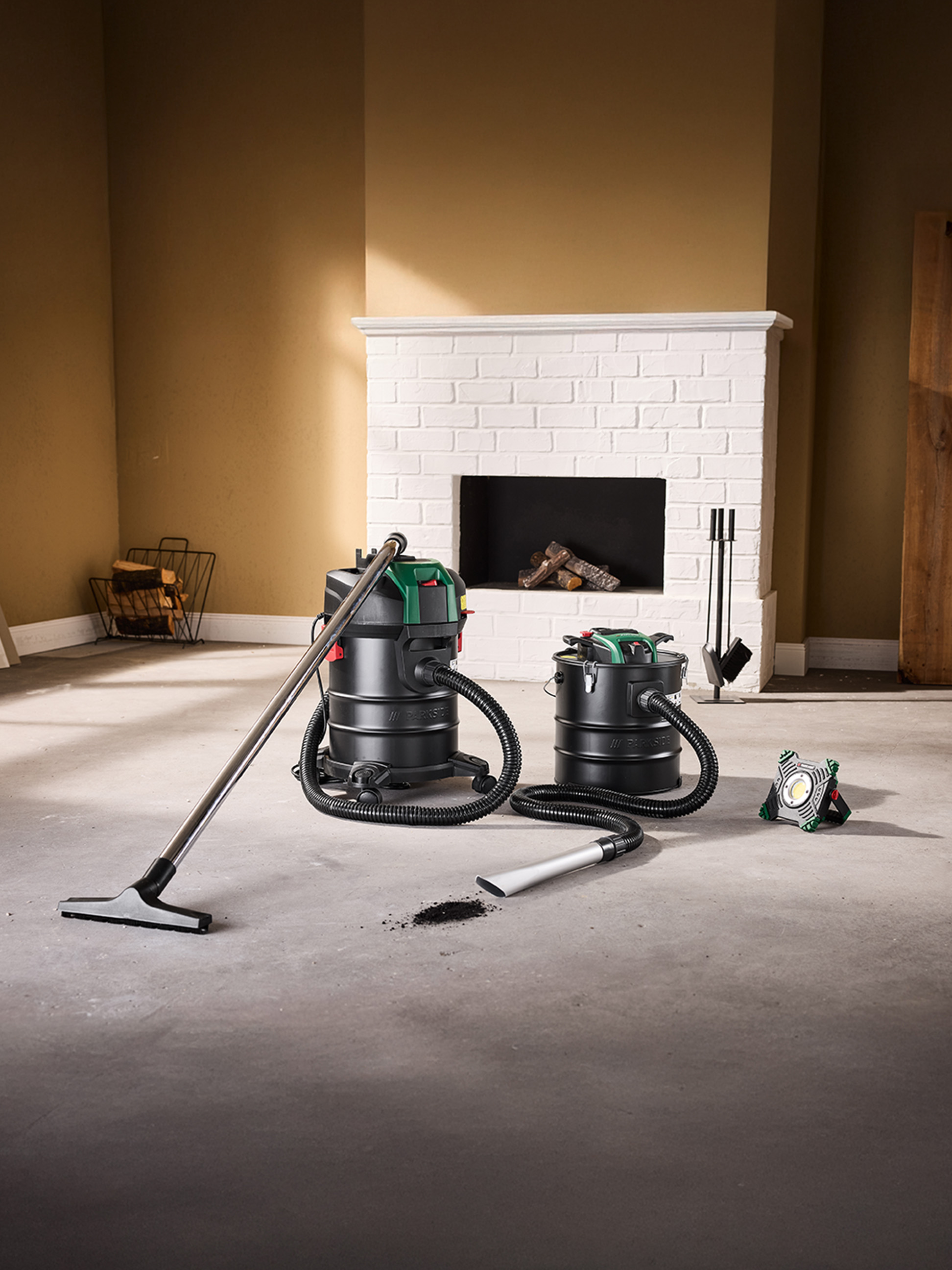 Deux aspirateurs à cendres et une lampe de travail sur un sol en béton devant une cheminée.