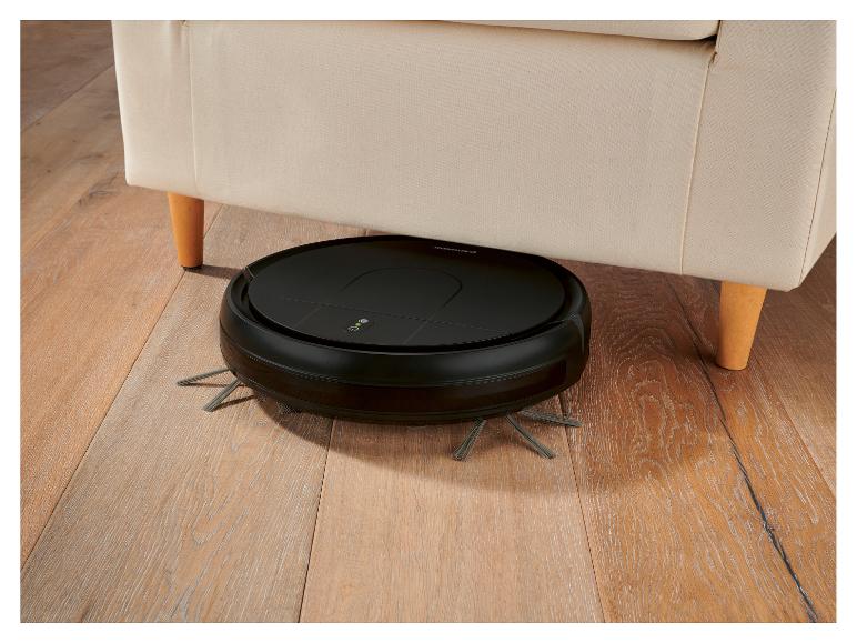 Aspirateur robot noir sous un canapé beige sur un parquet.