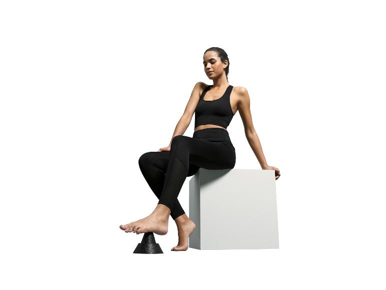 Femme en brassière de sport noire et leggings assise sur un cube blanc, pied sur un masseur noir.