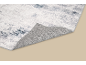 Tapis gris et blanc à motif abstrait, avec un coin replié, révélant son envers texturé.