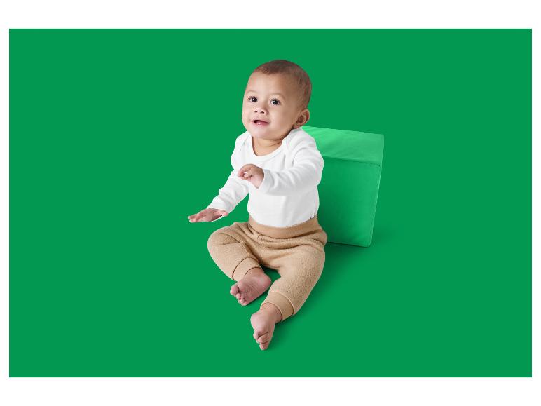 Bébé en body blanc à manches longues et pantalon beige, assis à côté d'un cube vert.