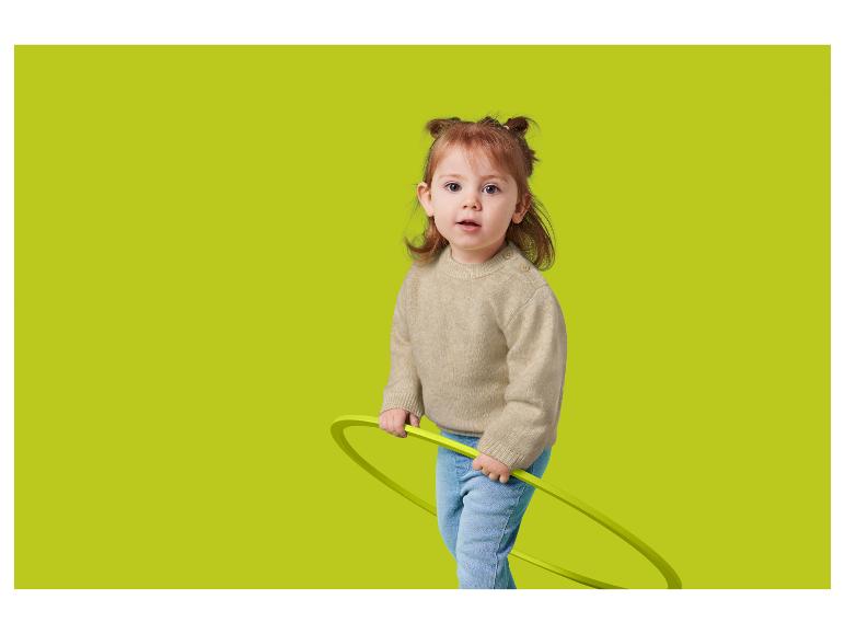 Un bambin aux cheveux roux, portant un pull beige et un jean, tenant un cerceau vert.