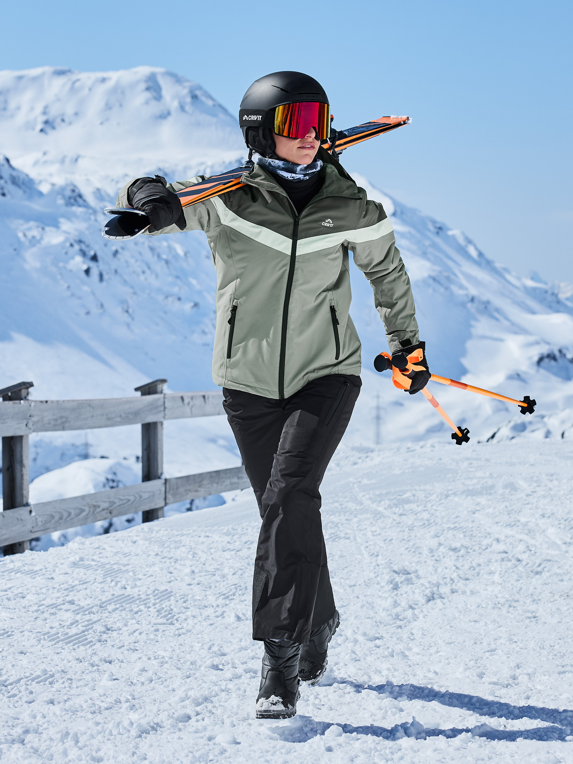 Femme en veste de ski, pantalon, casque, lunettes et skis sur l'épaule dans la neige.