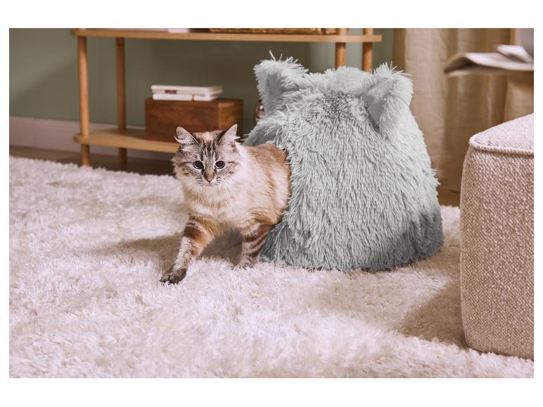 Un chat sort d'un lit douillet et moelleux sur un tapis blanc, à côté d'une étagère en bois.