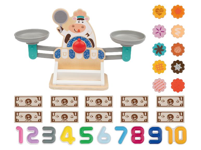 Une balance jouet en bois avec un design de vache, de la fausse monnaie, des chiffres et des formes de biscuits.