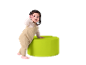 Bébé en salopette beige debout à côté d'un pouf vert.
