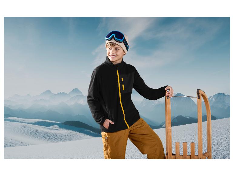 Un garçon avec une veste en polaire noire et des lunettes de ski s'appuie sur une luge en bois dans un paysage de montagne enneigé.