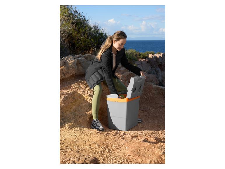 Femme ouvrant une glacière électrique grise et orange sur un terrain rocheux près de la mer.