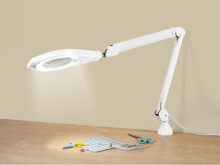 Lampe loupe blanche éclairant des fournitures de couture sur une table en bois.
