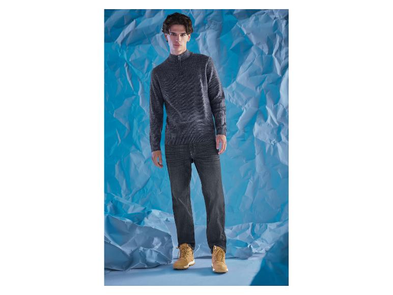 Un homme en pull gris et jean foncé devant un fond bleu.