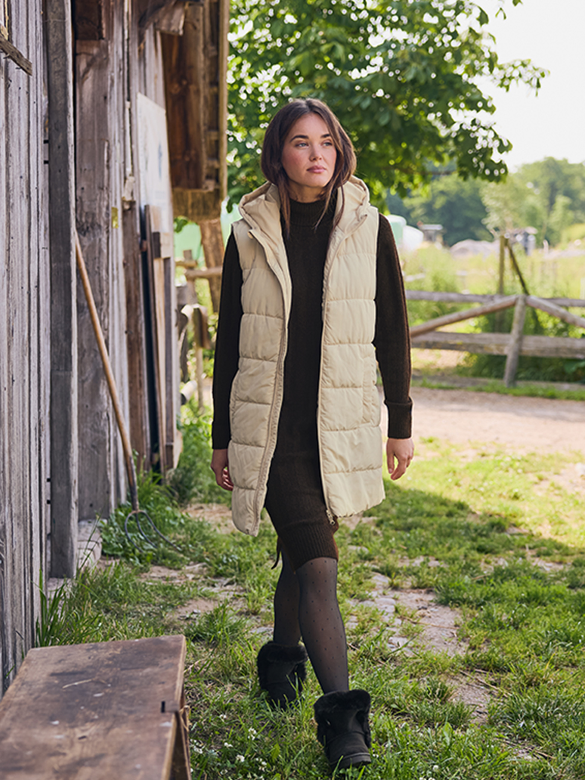 Femme en gilet beige et robe marron, marchant sur un chemin de campagne.