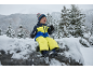 Enfant en combinaison de ski jaune et bleue, bonnet et gants, assis sur un rocher enneigé.