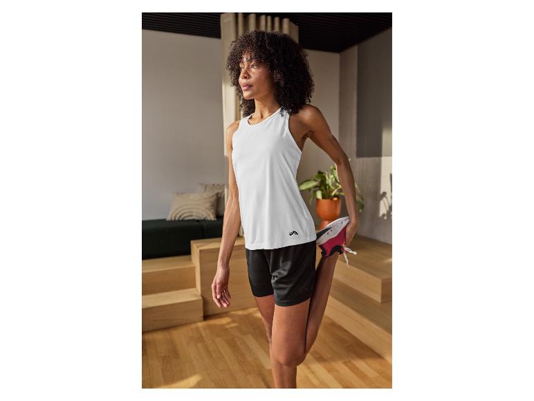 Femme en débardeur blanc et short noir, s'étirant dans une pièce lumineuse.