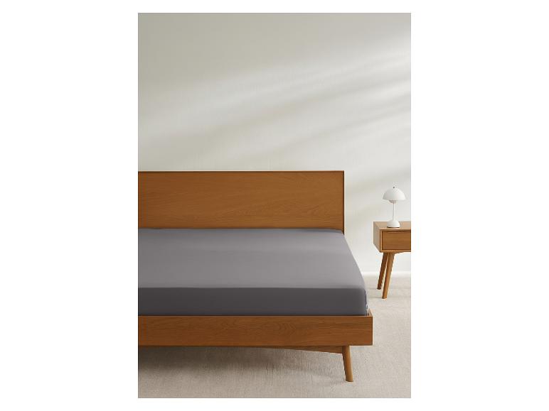 Lit avec cadre en bois et drap-housse gris, à côté d'une table de chevet avec lampe.