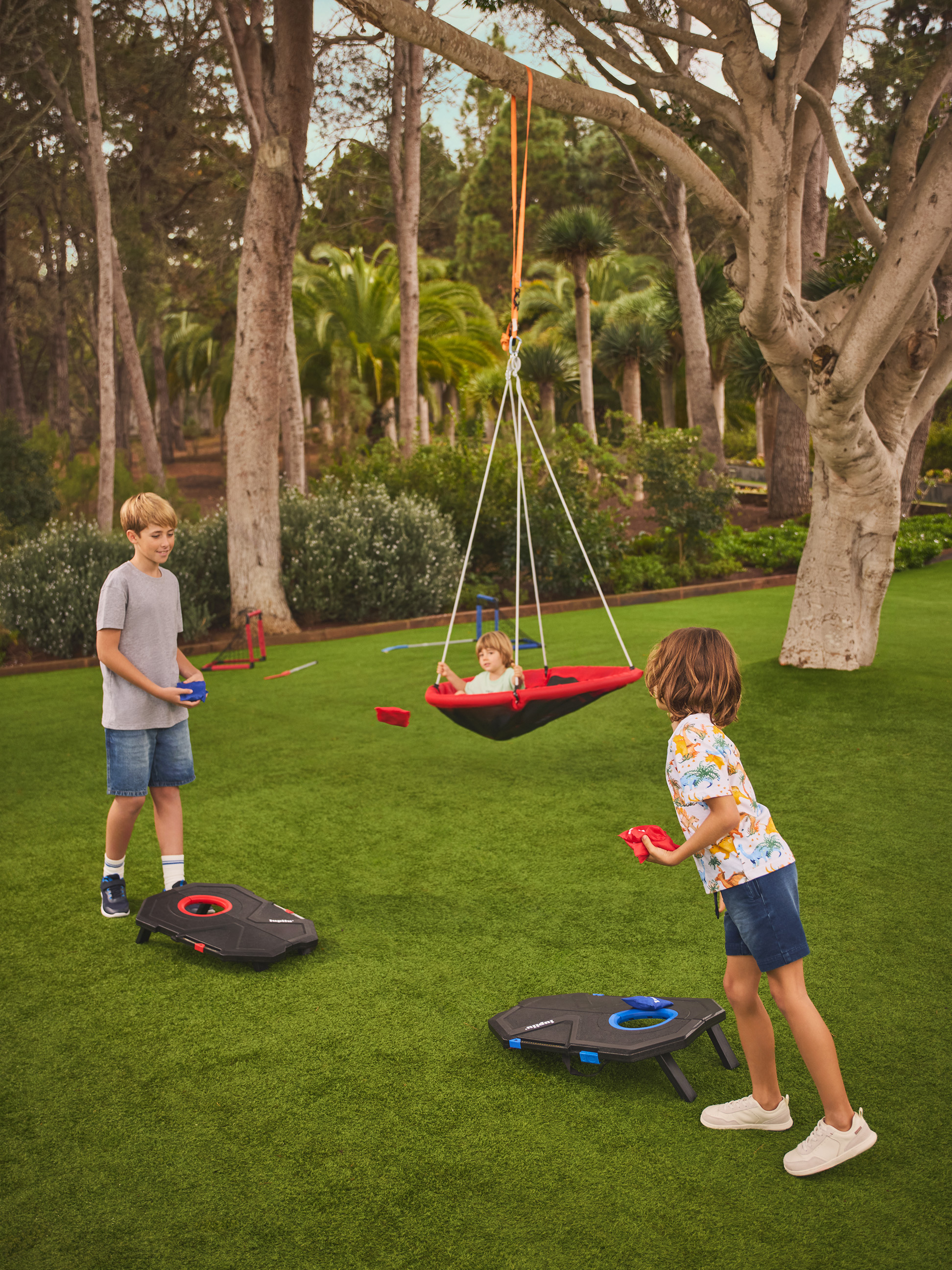 Deux enfants jouent au cornhole sur la pelouse, tandis qu'un troisième se balance dans une balançoire nid.