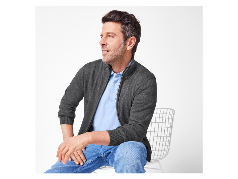 Un homme assis sur une chaise blanche porte un cardigan gris foncé zippé et un jean bleu.