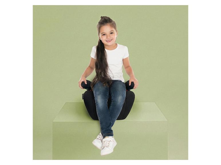 Fille souriante assise sur un rehausseur de voiture noir sur un piédestal vert.