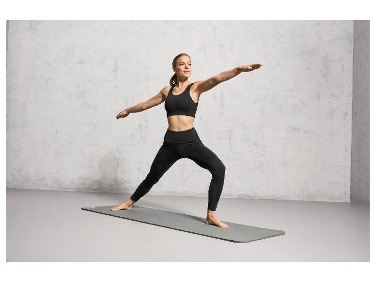 Une femme en brassière de sport et leggings fait du yoga sur un tapis gris.