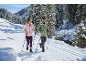 Couple en vestes et pantalons d'hiver avec bâtons sur une piste de ski en forêt enneigée.