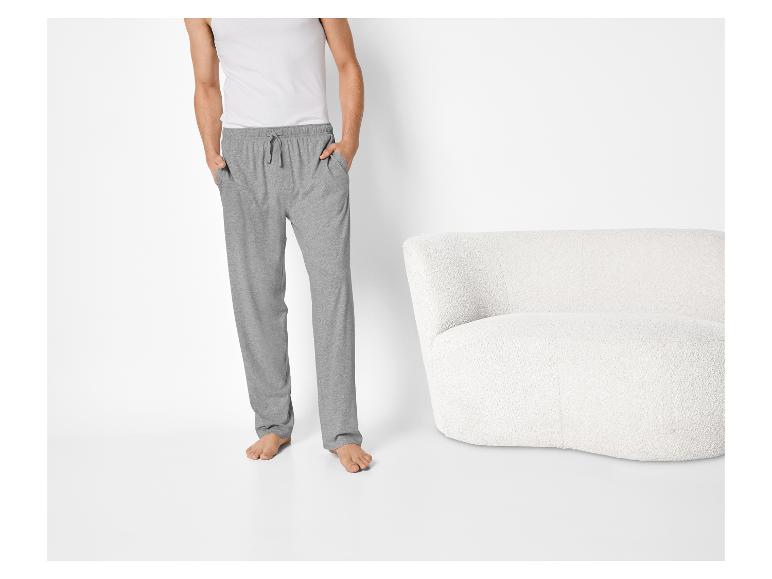 Homme en pantalon de survêtement gris et débardeur blanc, debout à côté d'un fauteuil blanc.