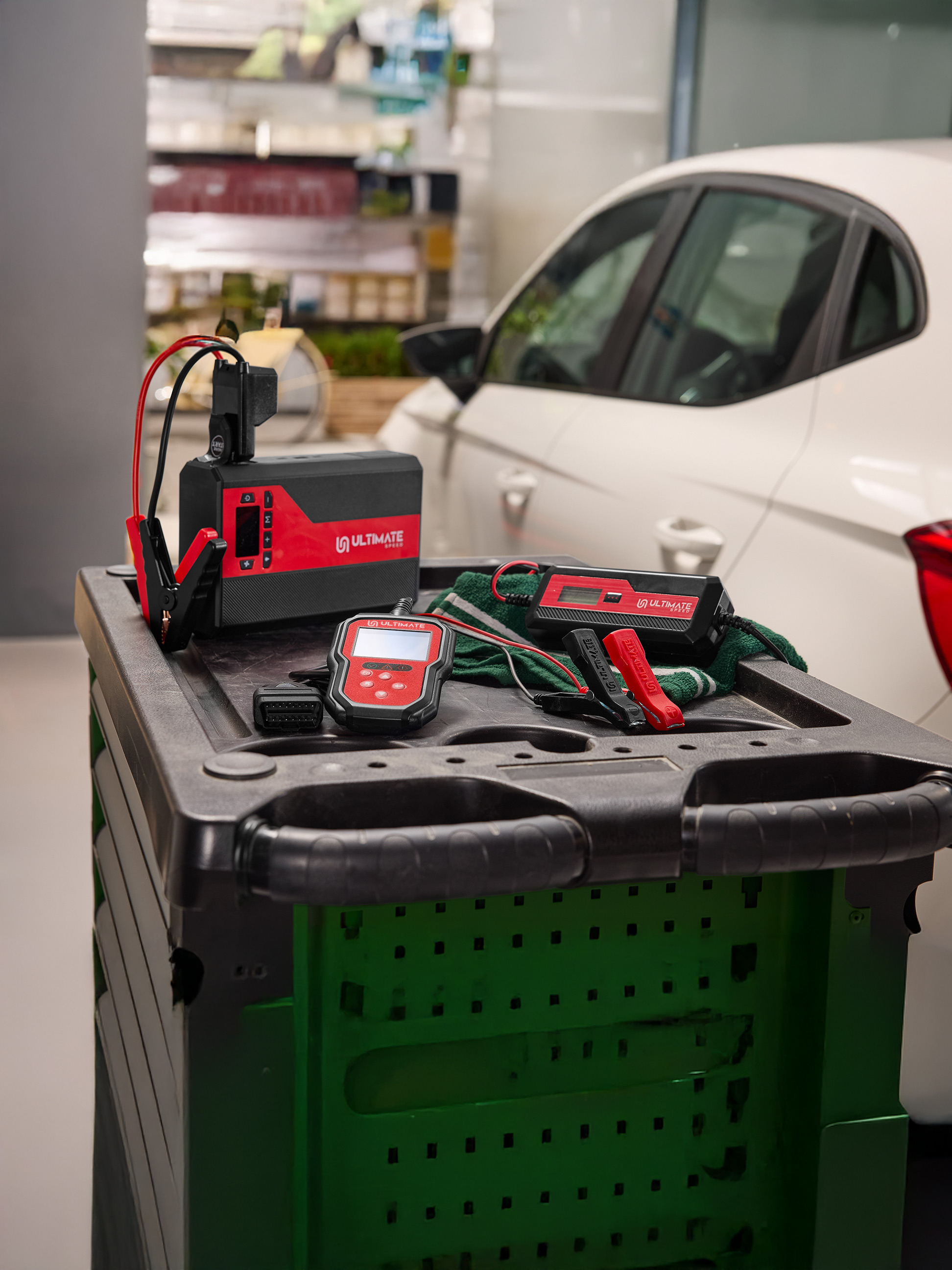 Chargeur de batterie de voiture et outils de diagnostic sur un chariot à côté d'une voiture blanche.