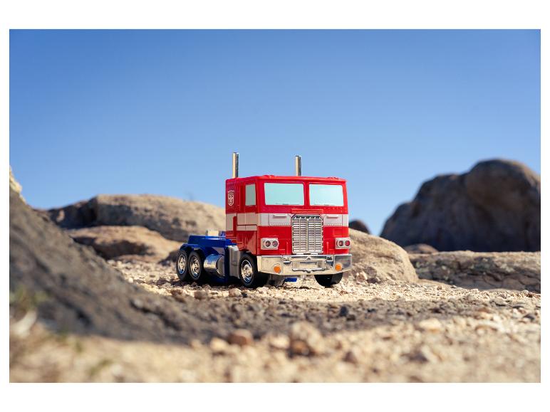 Un camion jouet Optimus Prime rouge et bleu sur un terrain rocheux et sablonneux sous un ciel clair.