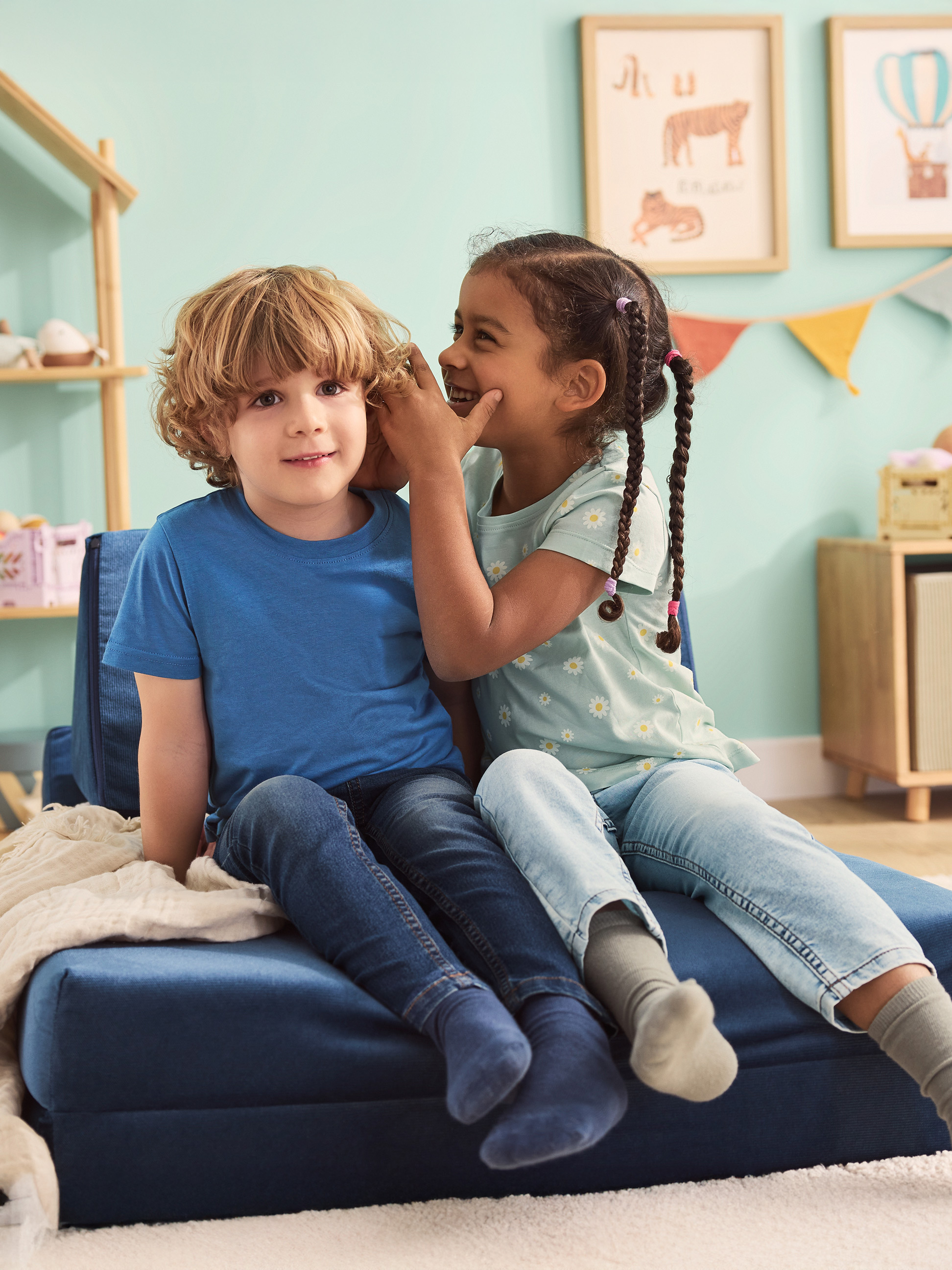 Deux enfants assis sur un canapé pliant bleu, l'un chuchotant à l'oreille de l'autre.