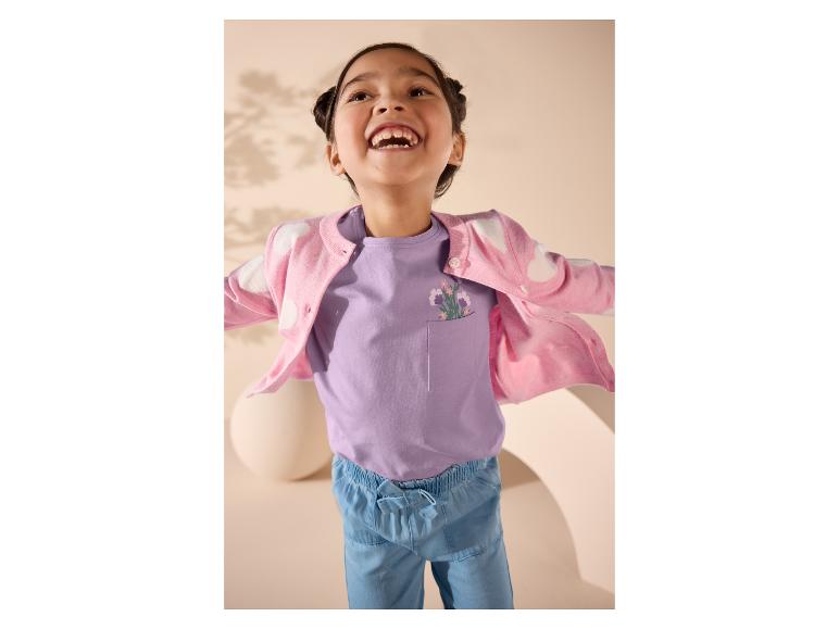 Fille souriante en cardigan rose, t-shirt violet à poche et pantalon bleu.
