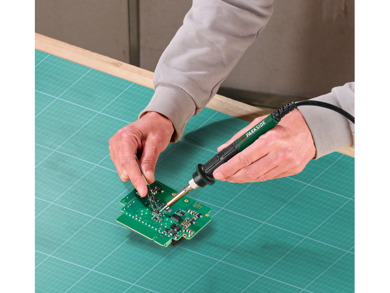 Mains soudant une carte de circuit imprimé avec un fer à souder sur un tapis de découpe vert.