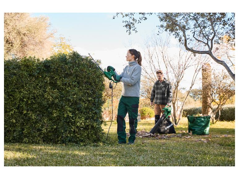 Une femme utilise une taille-haie Parkside tandis qu'un homme utilise un souffleur à feuilles dans un jardin.