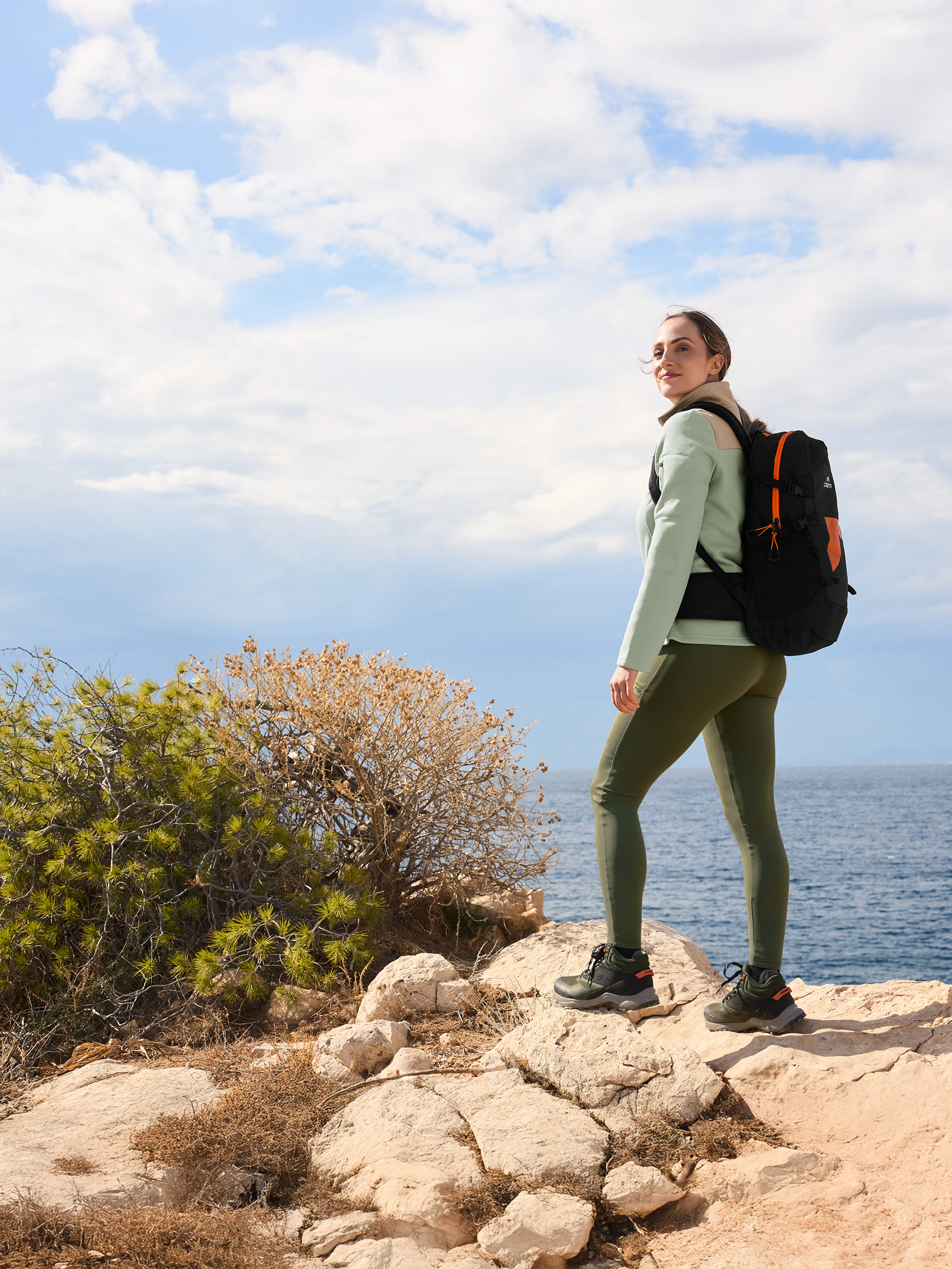 Femme en tenue de randonnée avec sac à dos sur un rocher près de la mer.