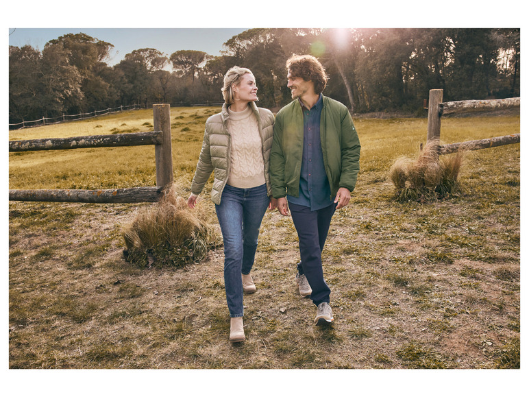 Un couple se promène main dans la main dans un champ, ils portent des vestes d'hiver et des jeans.