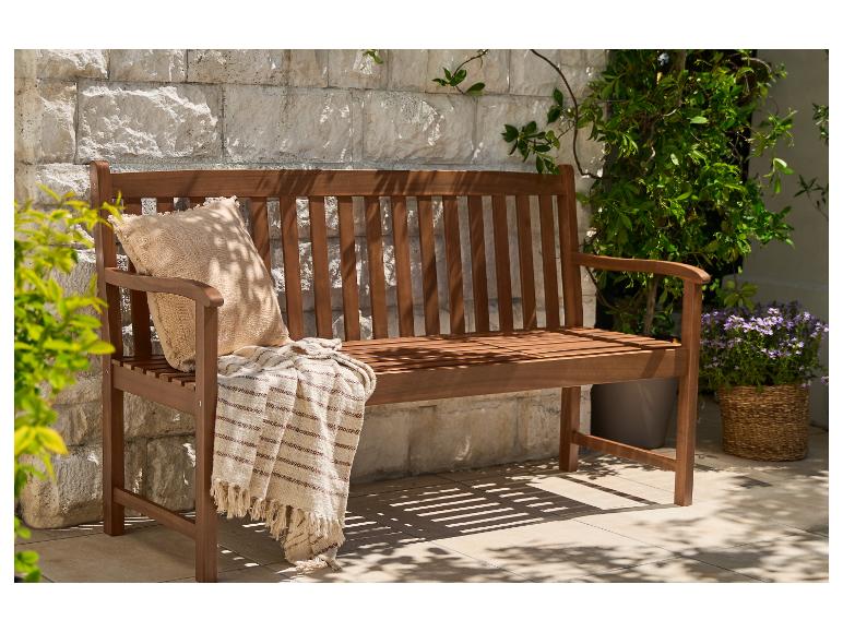 Banc de jardin en bois avec coussin et plaid, devant un mur de pierre avec des plantes.