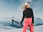 Une femme en pantalon de ski rouge et un pull noir tenant des bâtons de ski avec des montagnes en arrière-plan.