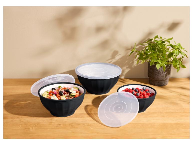 Trois saladiers noirs avec couvercles transparents, remplis de salade et de baies, sur une table en bois.