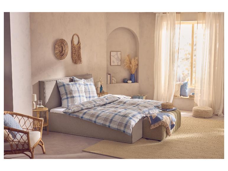 Chambre avec un lit beige, une parure de lit à carreaux bleu et blanc, un fauteuil en osier et des objets décoratifs.