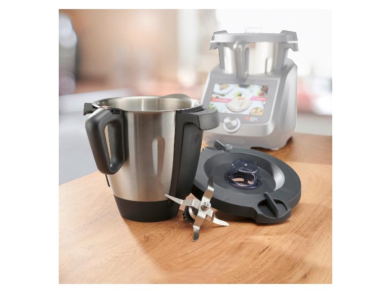 Bol mélangeur en acier inoxydable avec lames et couvercle, avec robot culinaire en arrière-plan.