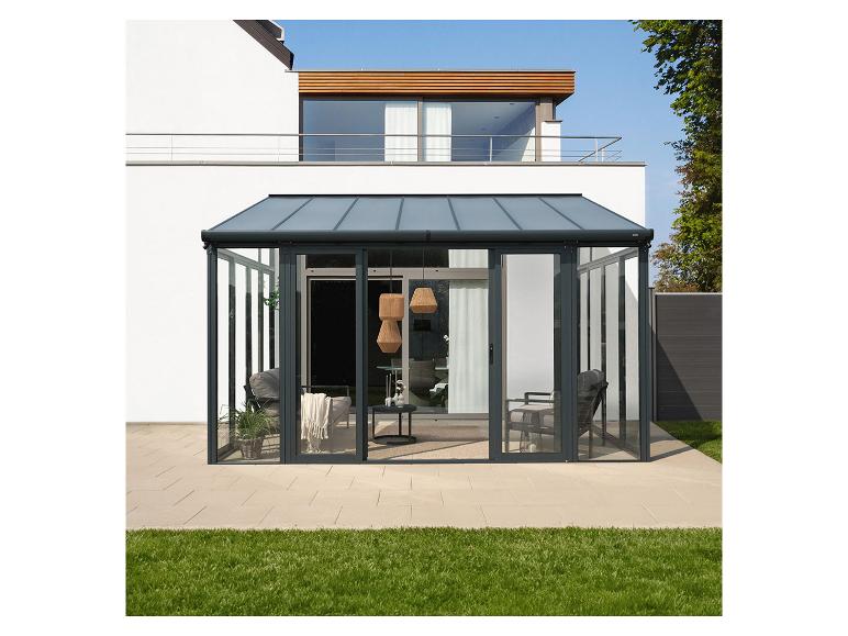 Une véranda en verre avec des cadres gris foncé et des meubles, devant une maison et une pelouse.