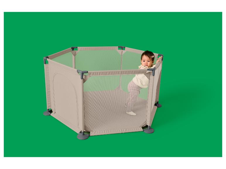 Bébé dans un parc hexagonal beige avec des parois en maille, debout et appuyé sur le bord.