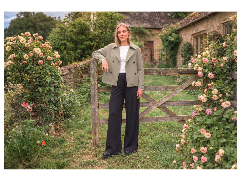 Femme en veste verte et pantalon noir large près d'une barrière en bois avec des roses.