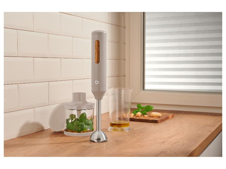 Mixeur plongeant avec basilic, huile d'olive et fromage sur un comptoir en bois dans une cuisine.