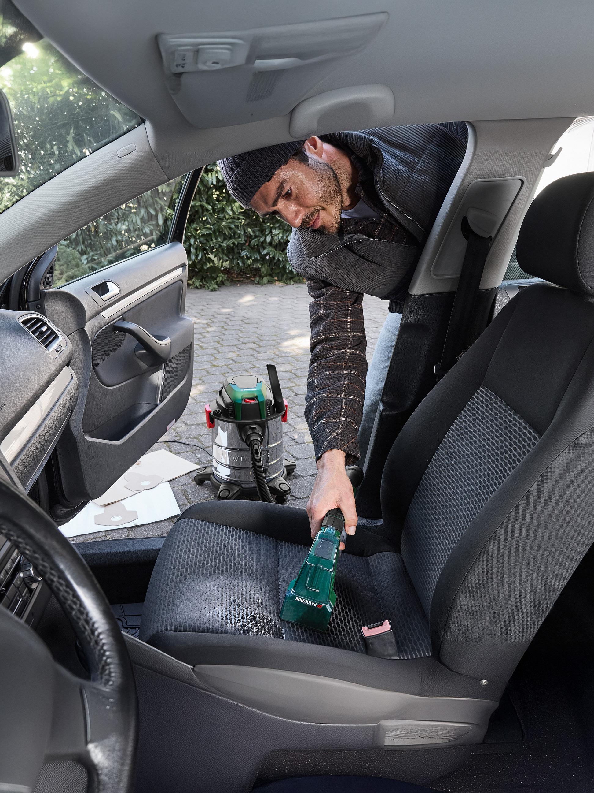 Homme nettoyant un siège de voiture avec un aspirateur à main Parkside, avec un aspirateur eau et poussière en arrière-plan.